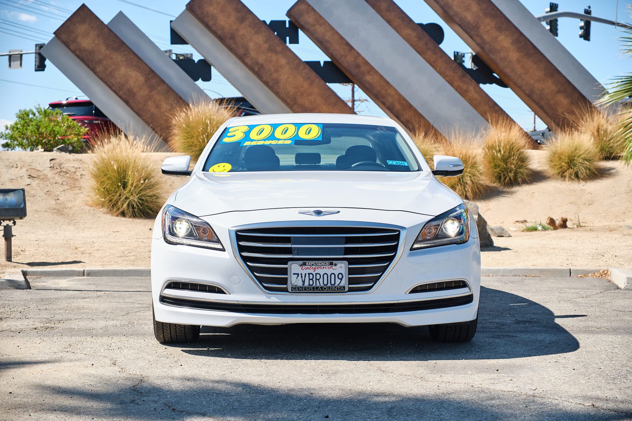 A front view of a white Hyundai Genesis luxury sedan with artistic angular sculptures in the background.