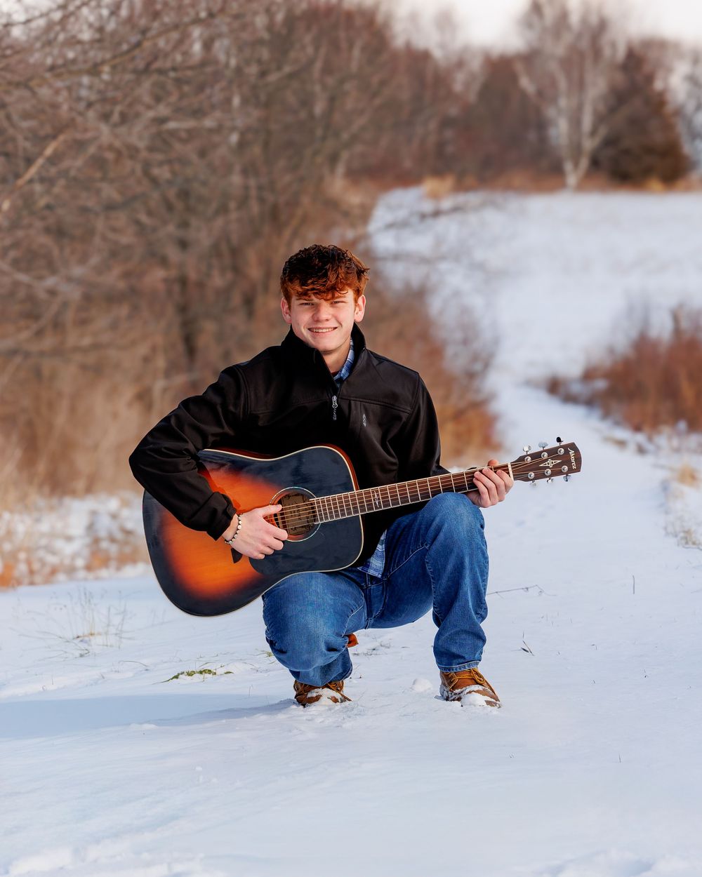 Young man crouching in snow holding acoustic guitar, wearing black jacket and jeans, smiling outdoors in winter landscape.