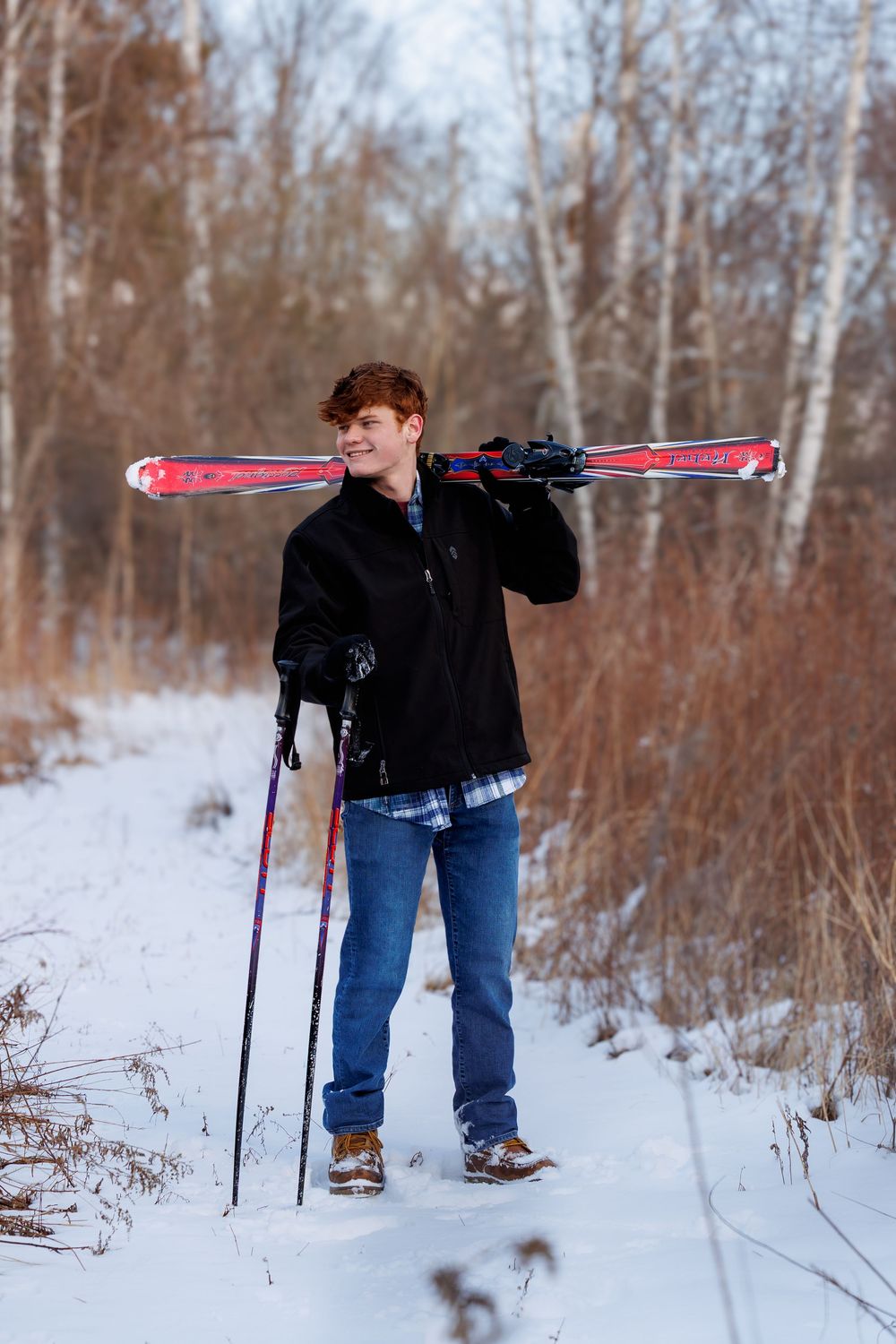 Young man carrying red skis on his shoulder while standing on a snowy trail surrounded by winter trees.