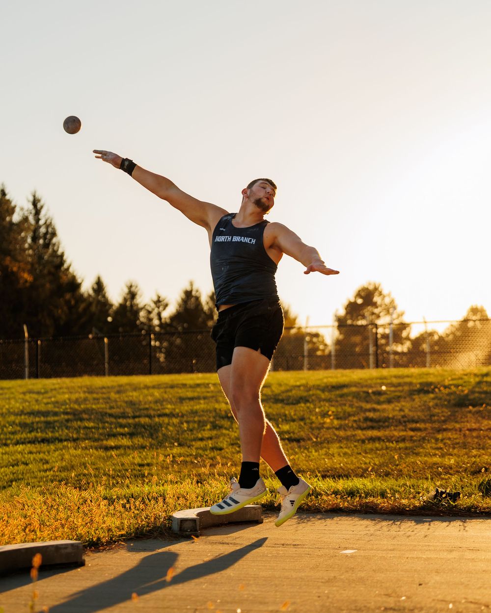 A dancer performs a graceful leap in a grassy field at sunset, casting a long shadow on the ground.