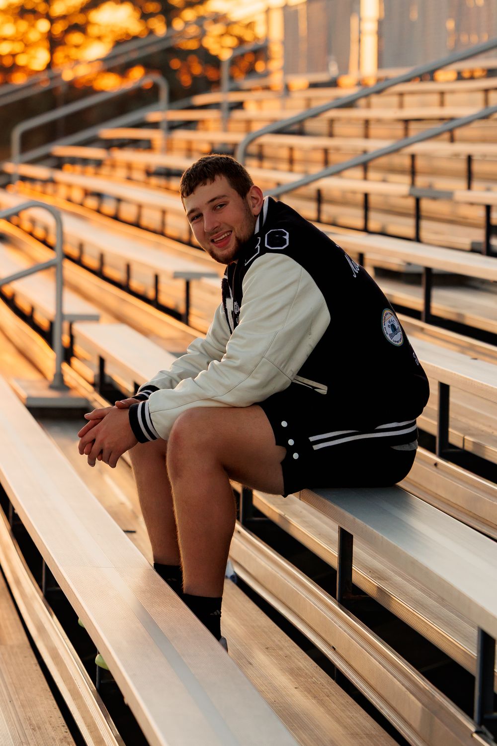 Someone wearing a varsity letterman jacket sits on bleacher seats at sunset with warm golden lighting.