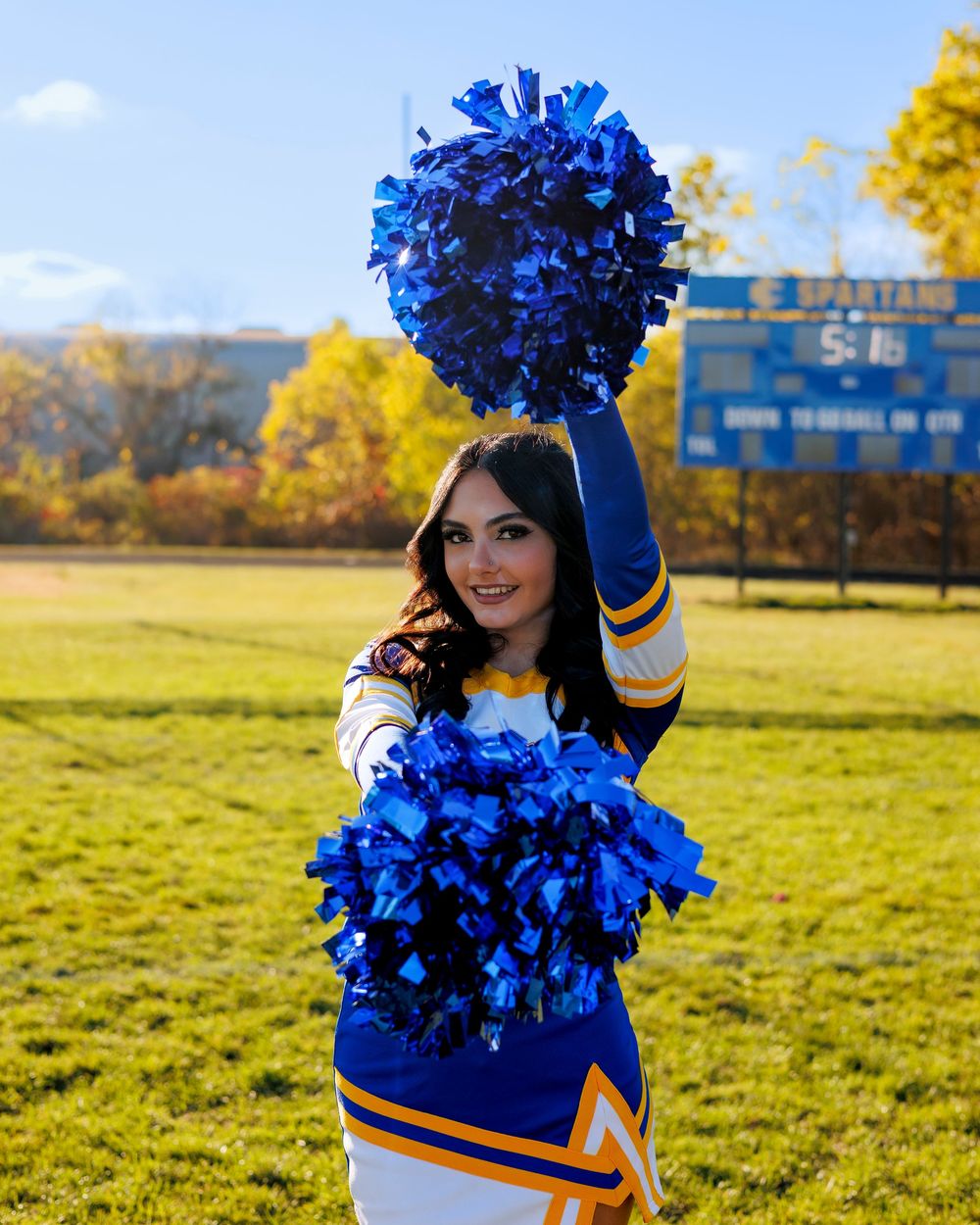 A cheerleader in blue and white uniform holds up pom poms on a sunny autumn day with yellow trees in the background.
