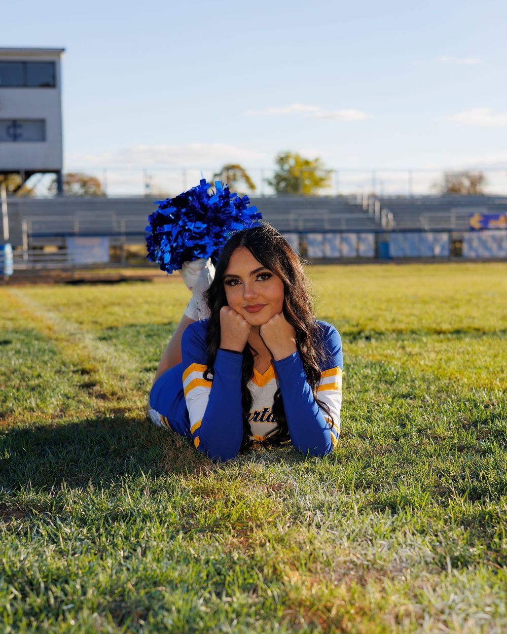 A person wearing a blue floral crown and blue outfit lies on green grass during a sunny day at a sports field.