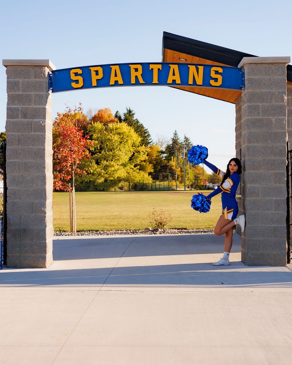 A cheerleader performs splits between brick pillars with a blue 'Spartans' banner and autumn trees in background.