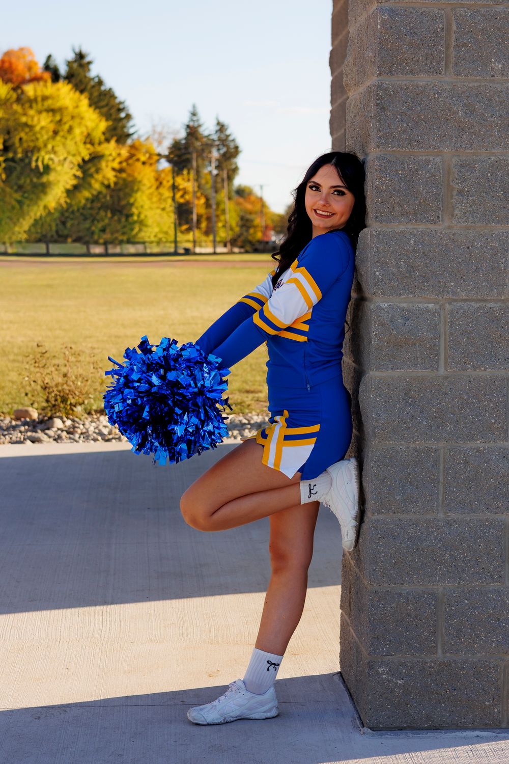Cheerleader in blue and gold uniform performs dynamic poses against brick wall and autumn background.