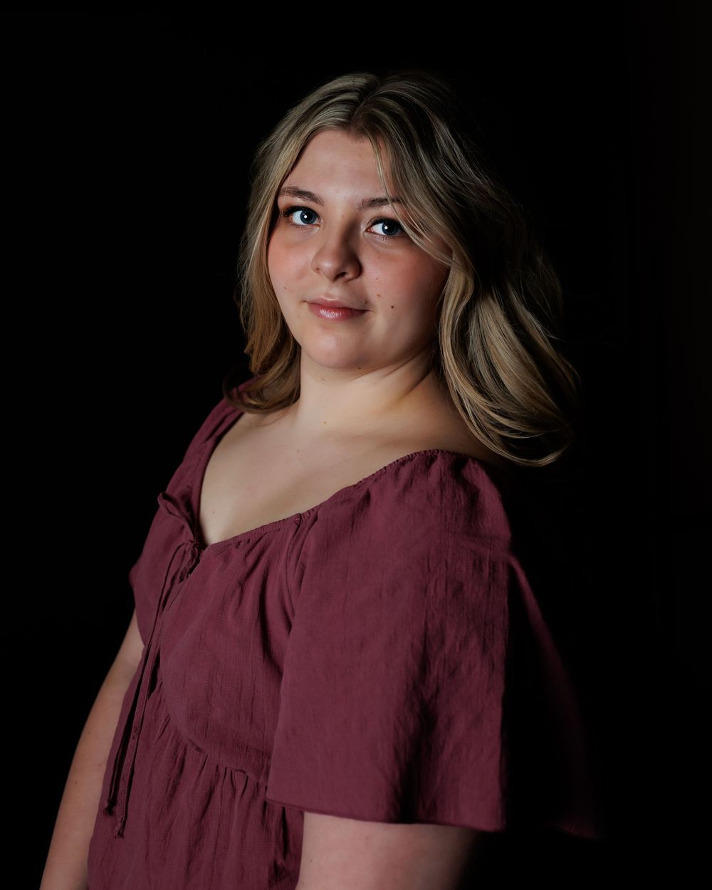 Studio portrait of someone wearing a burgundy peasant top dress against a dark background.