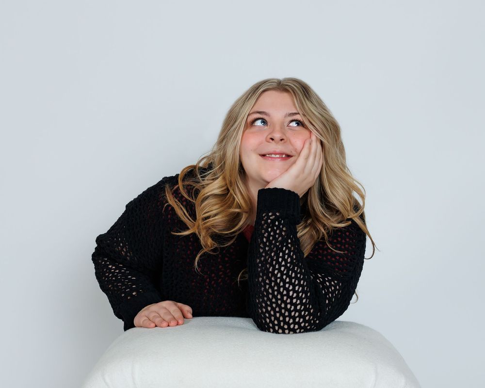 Person with blonde wavy hair wearing a black sweater leans on white table while looking up thoughtfully.