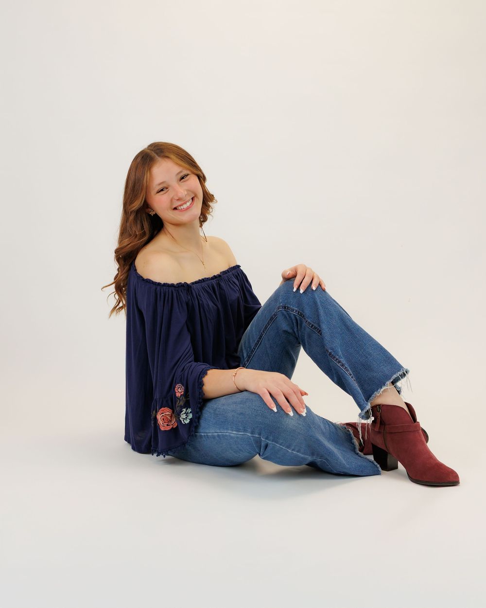 A person in a navy off-shoulder top with embroidery detail lounges casually in blue jeans and burgundy ankle boots.