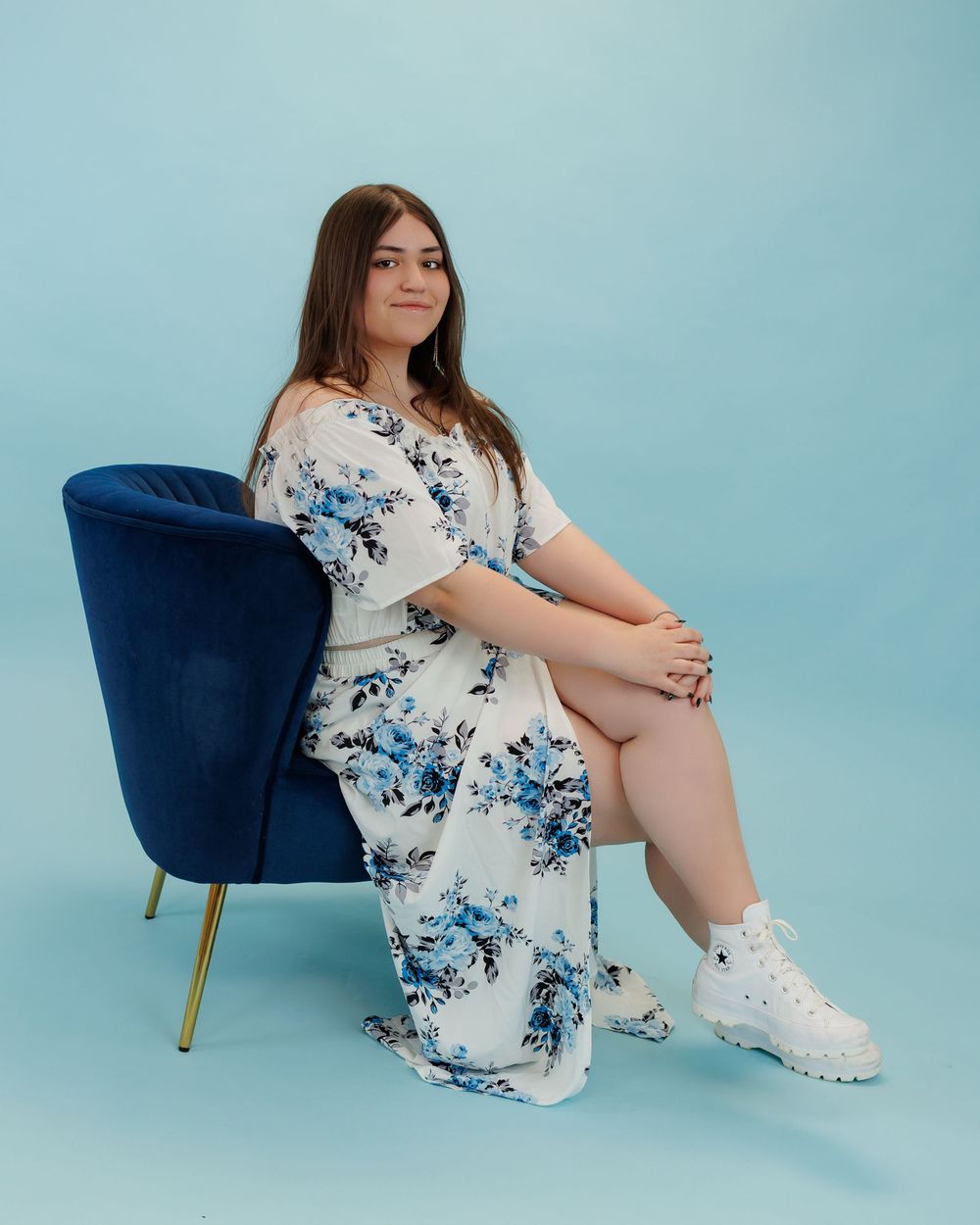 A person in a floral white and blue maxi dress sits on a navy velvet chair against a light blue backdrop.