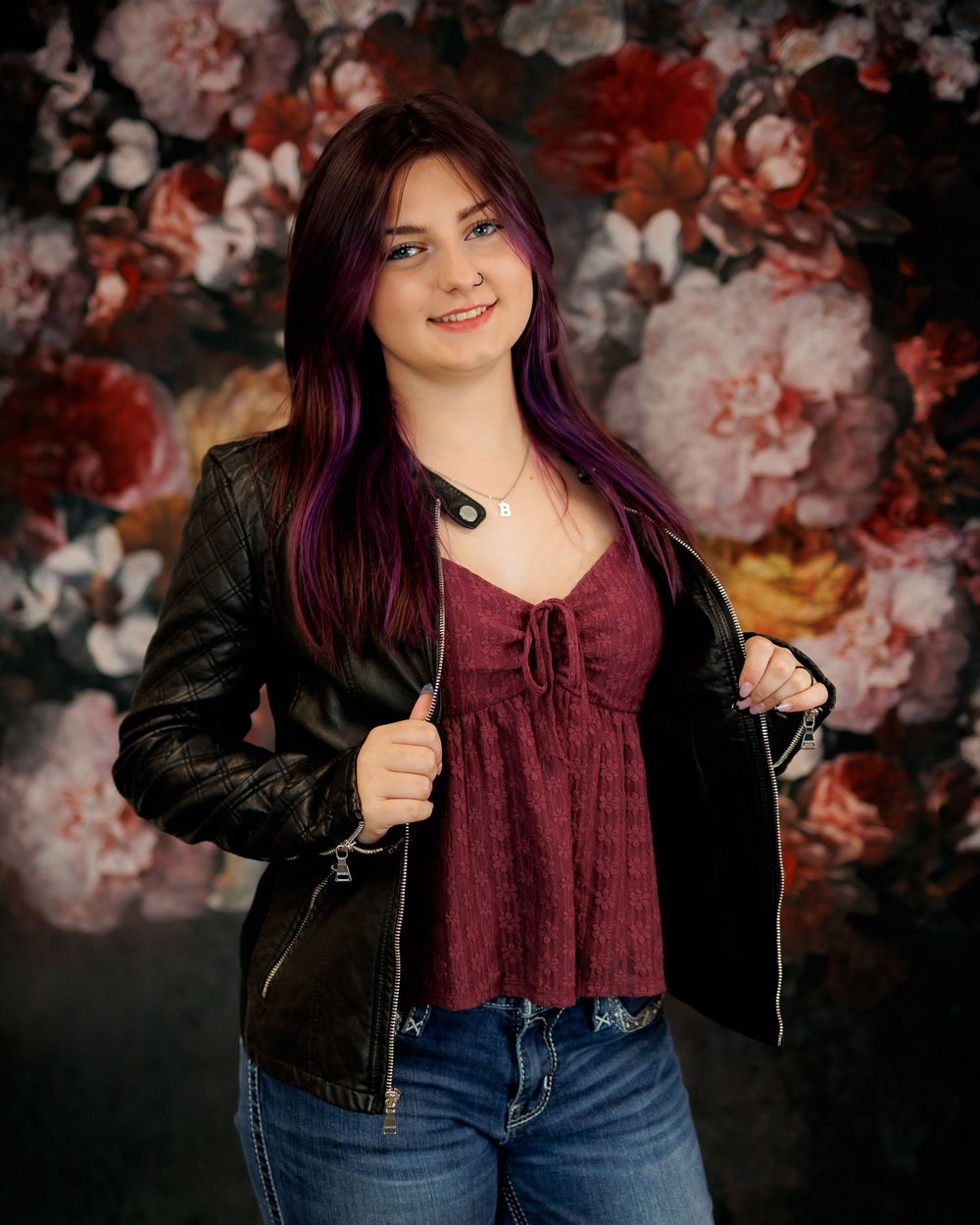 A person in a black jacket and burgundy top poses against a floral backdrop in a photo studio setting.