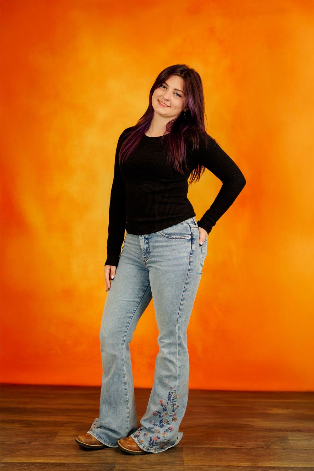 A person in a black top and embroidered jeans poses against an orange background on a wooden floor.
