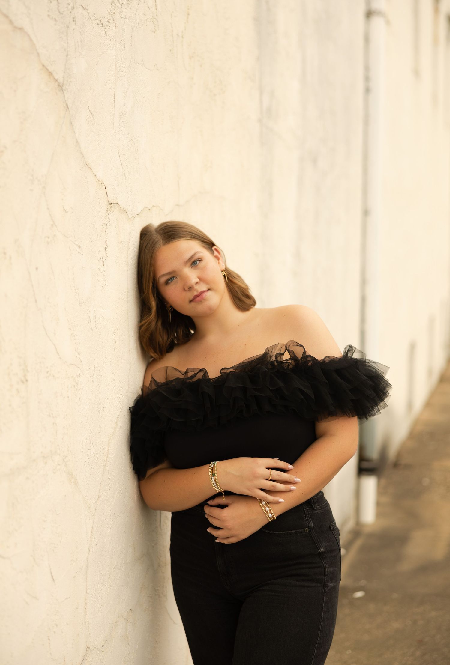A curvy model in an elegant black off-shoulder dress poses against a white wall in natural lighting.