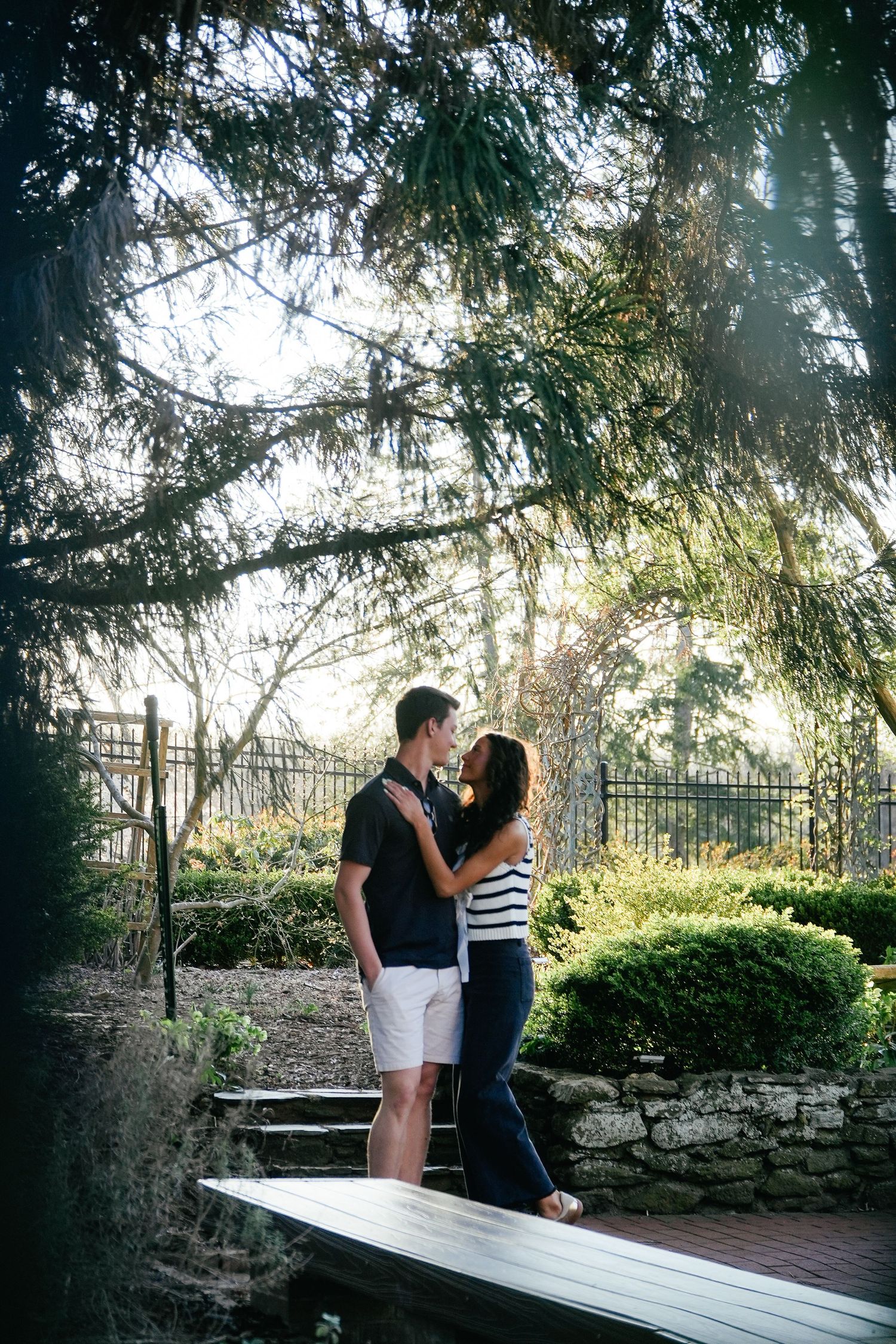 A couple shares a romantic moment on a wooden deck surrounded by lush greenery and stone walls in a garden setting.