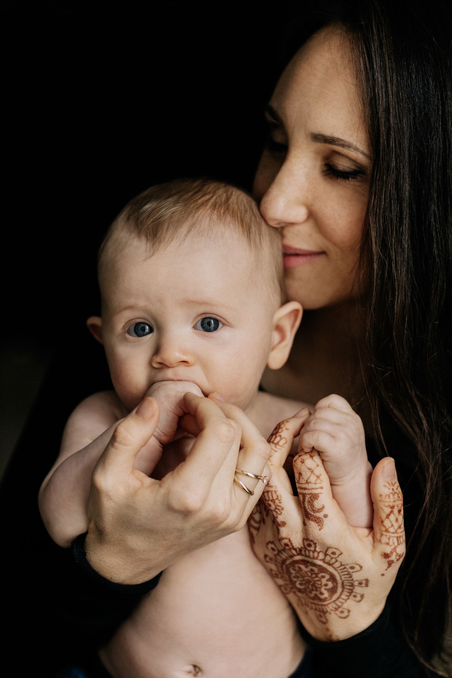zärtliches Motherhood-Portrait einer Mutter, die ihr nacktes Kind auf den Kopf küsst, welches an seiner Hand lutscht. People-und Lifestyle
