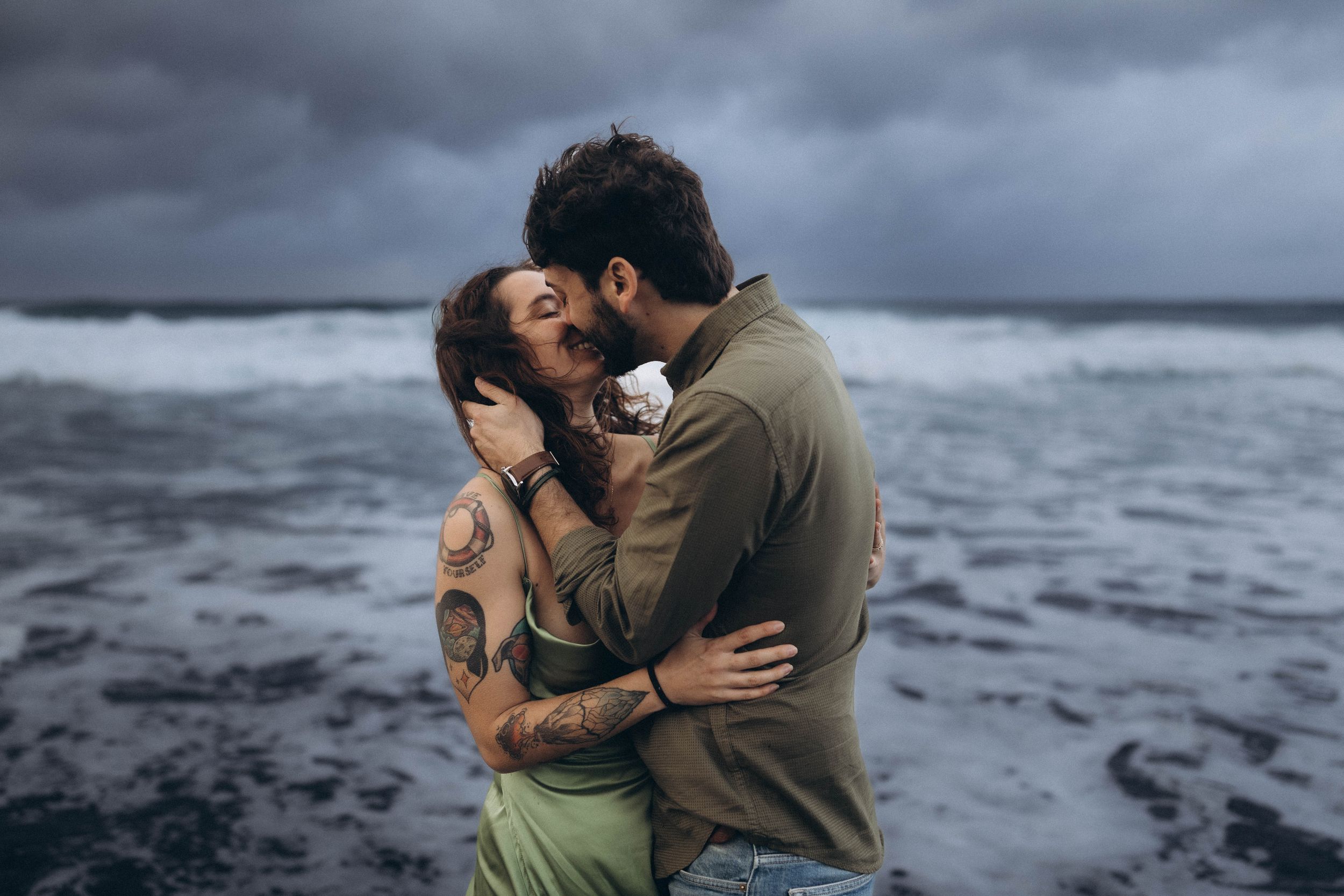 A couple embraces in a passionate kiss against a dramatic stormy ocean backdrop with crashing waves.