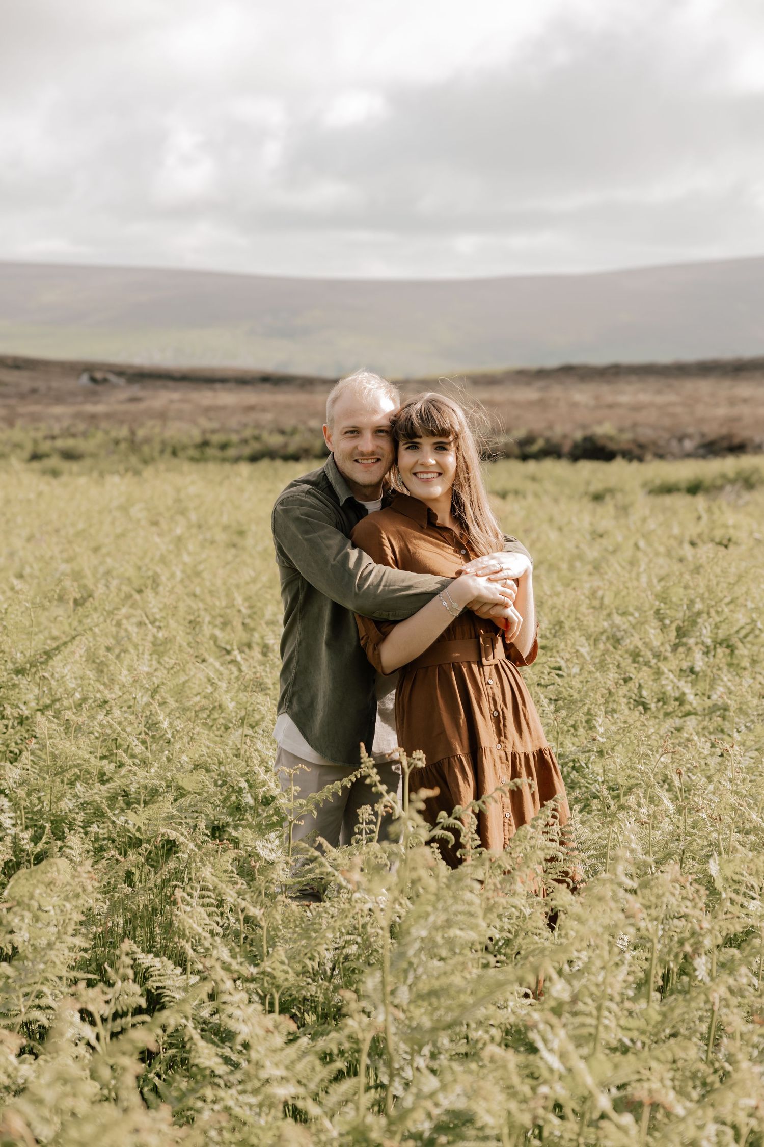 Two people embrace while standing together in a grassy meadow with rolling hills in the background under cloudy skies.