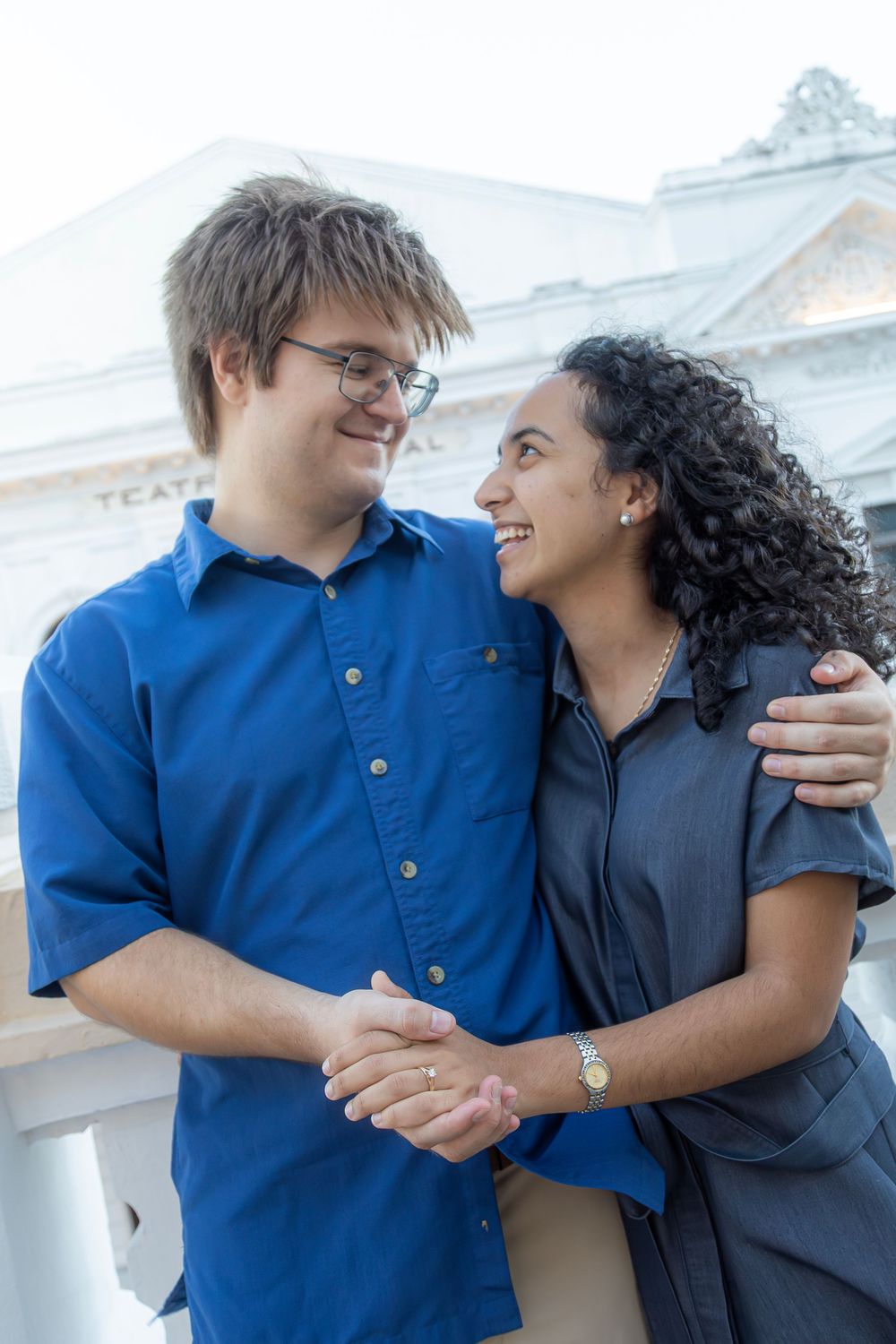 A happy couple in blue and gray outfits embrace while standing outside a white building with architectural details.