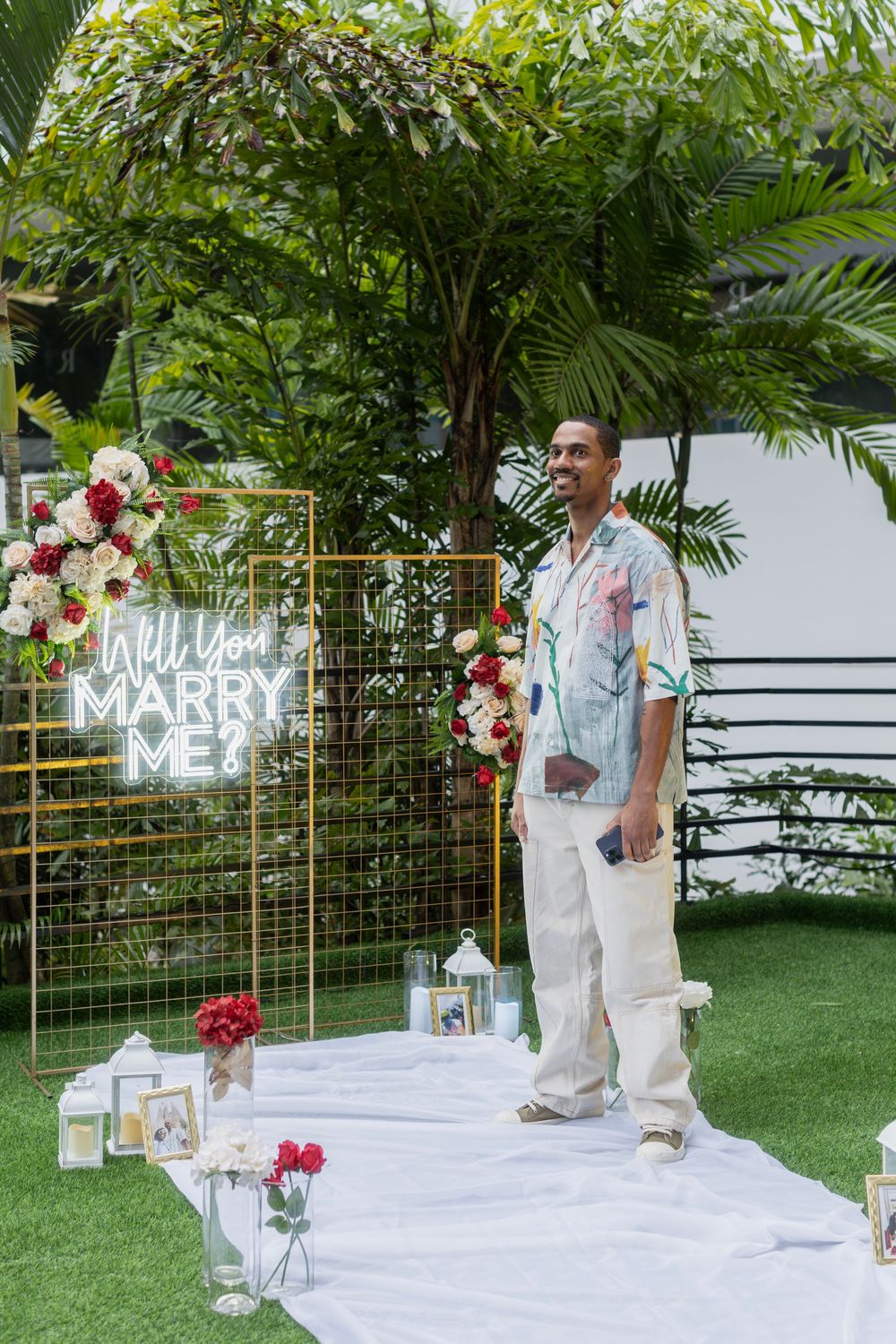 Garden proposal setting with neon 'Marry Me' sign and floral decorations against tropical foliage.