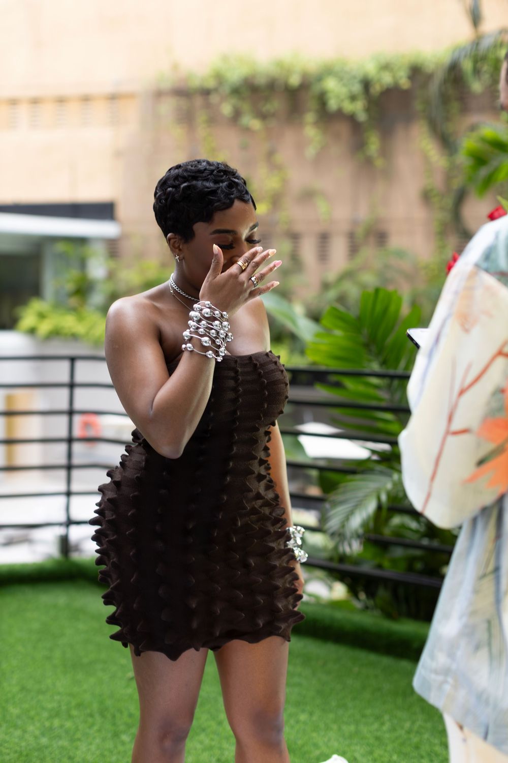 Woman in a textured black dress covering her mouth in surprise while standing outdoors.