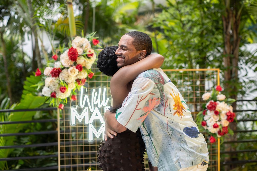 Newly engaged couple embracing in front of floral decorations and neon signage.
