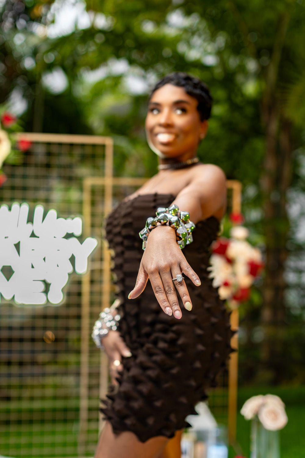 Woman showing engagement ring while smiling in front of decorative backdrop with flowers.