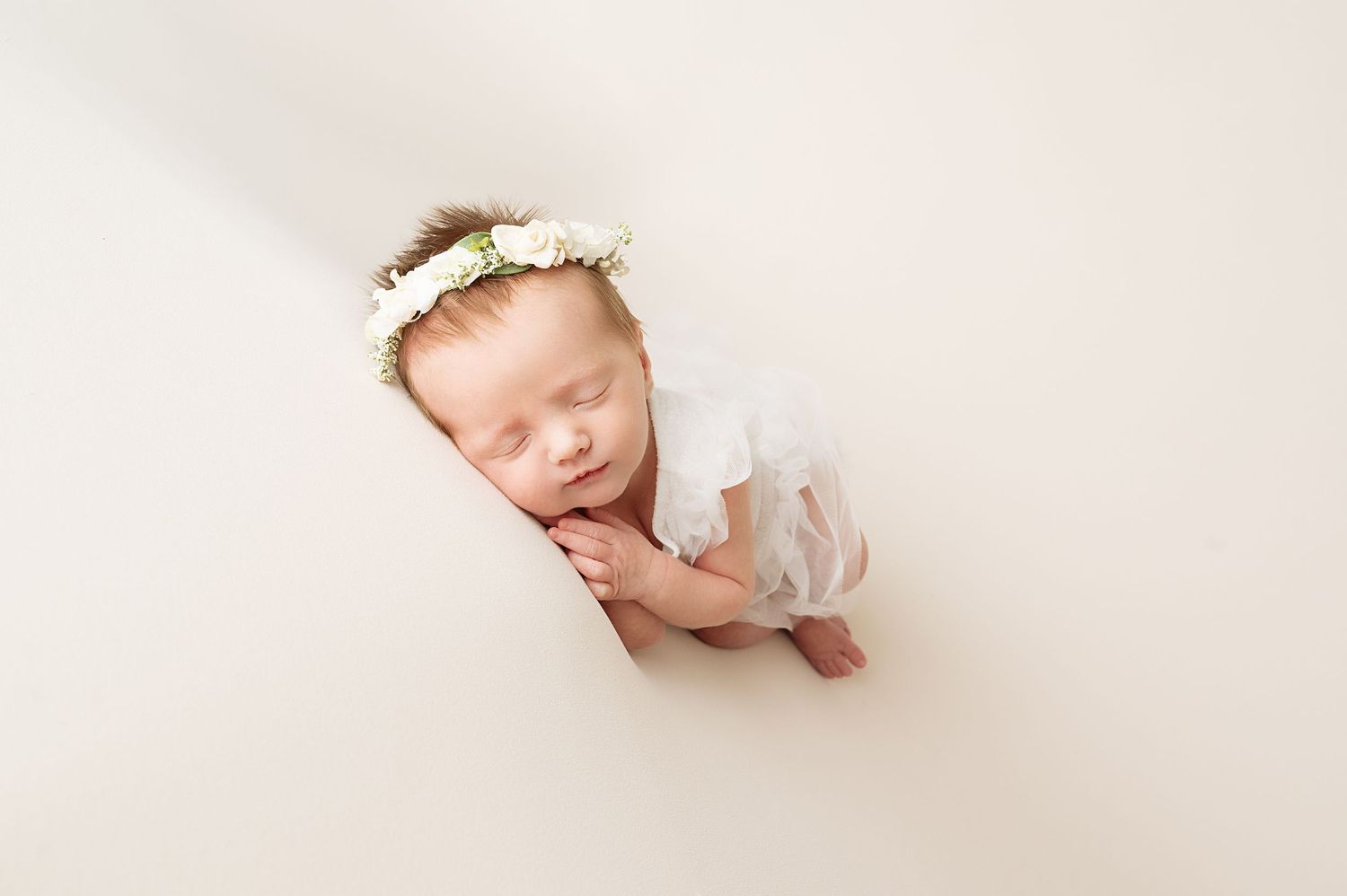 Sleeping newborn swaddled in white fabric wearing delicate floral headband on cream background.
