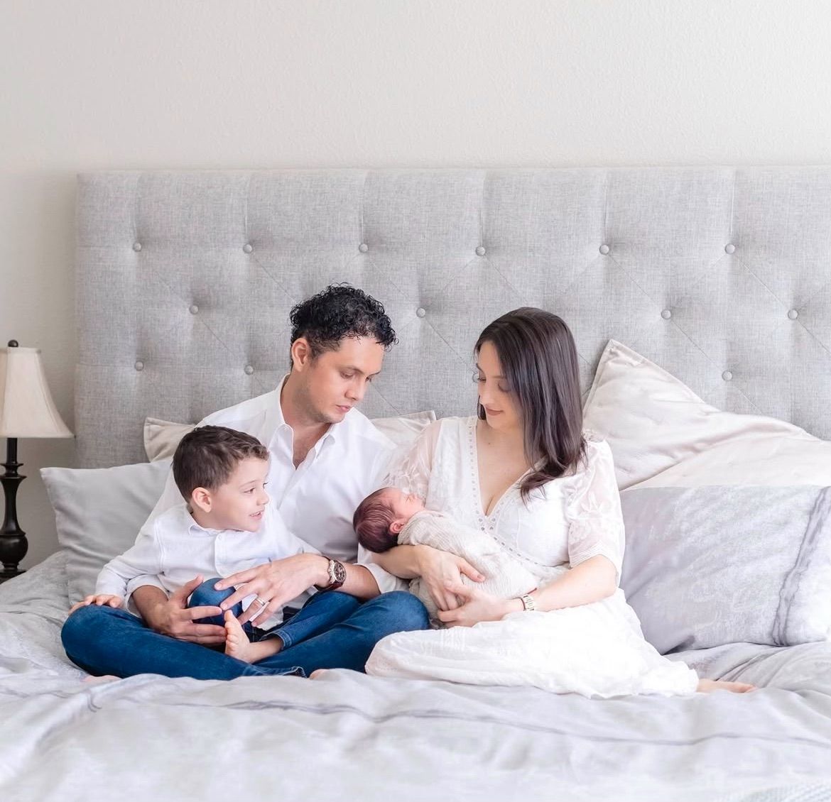 Family relaxing together on gray upholstered bed with newborn baby.
