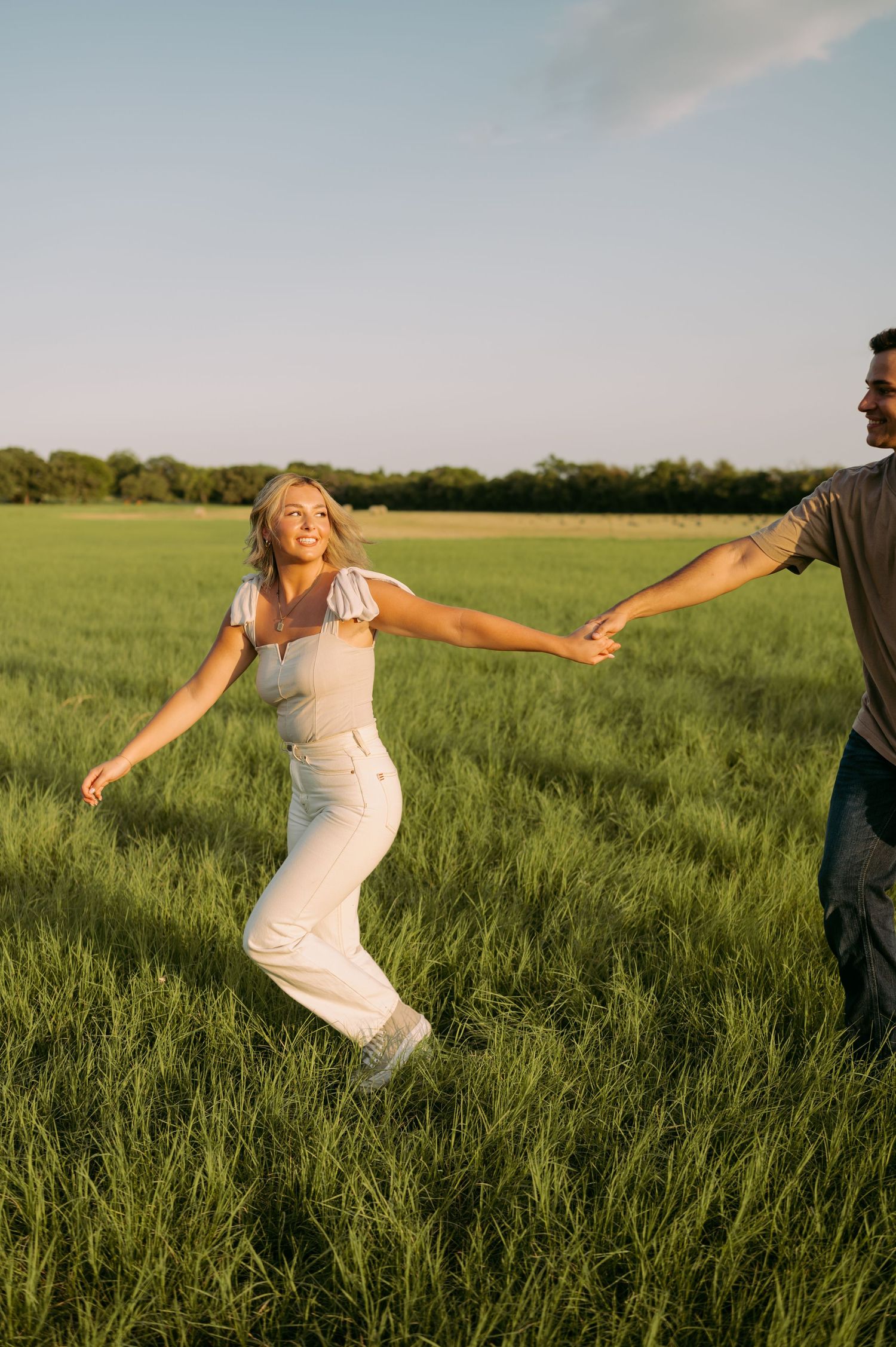 A couple holds hands while dancing together in a sunlit green meadow during golden hour.