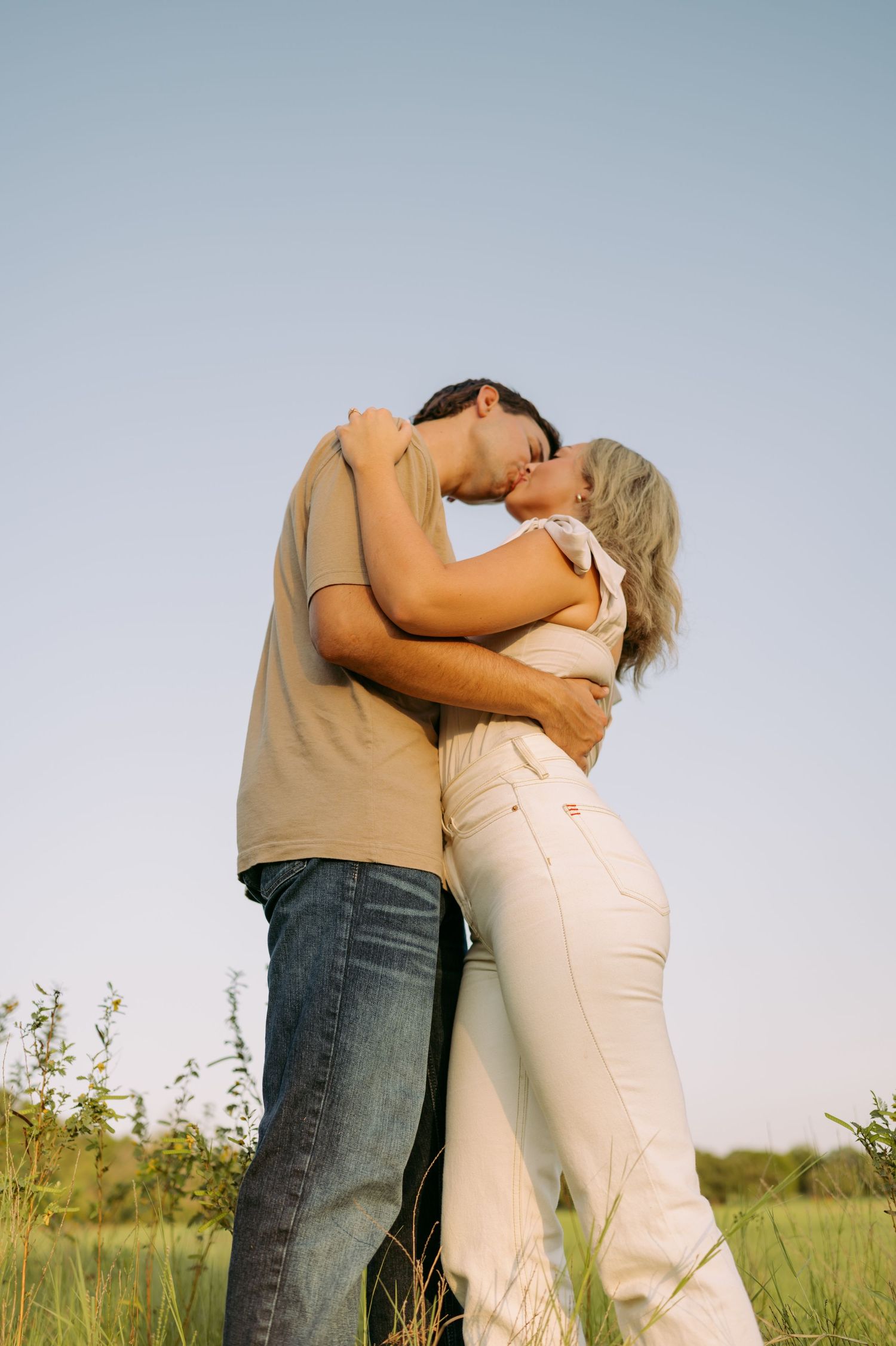 An intimate moment captured between a couple in casual attire standing in a grassy field at sunset.