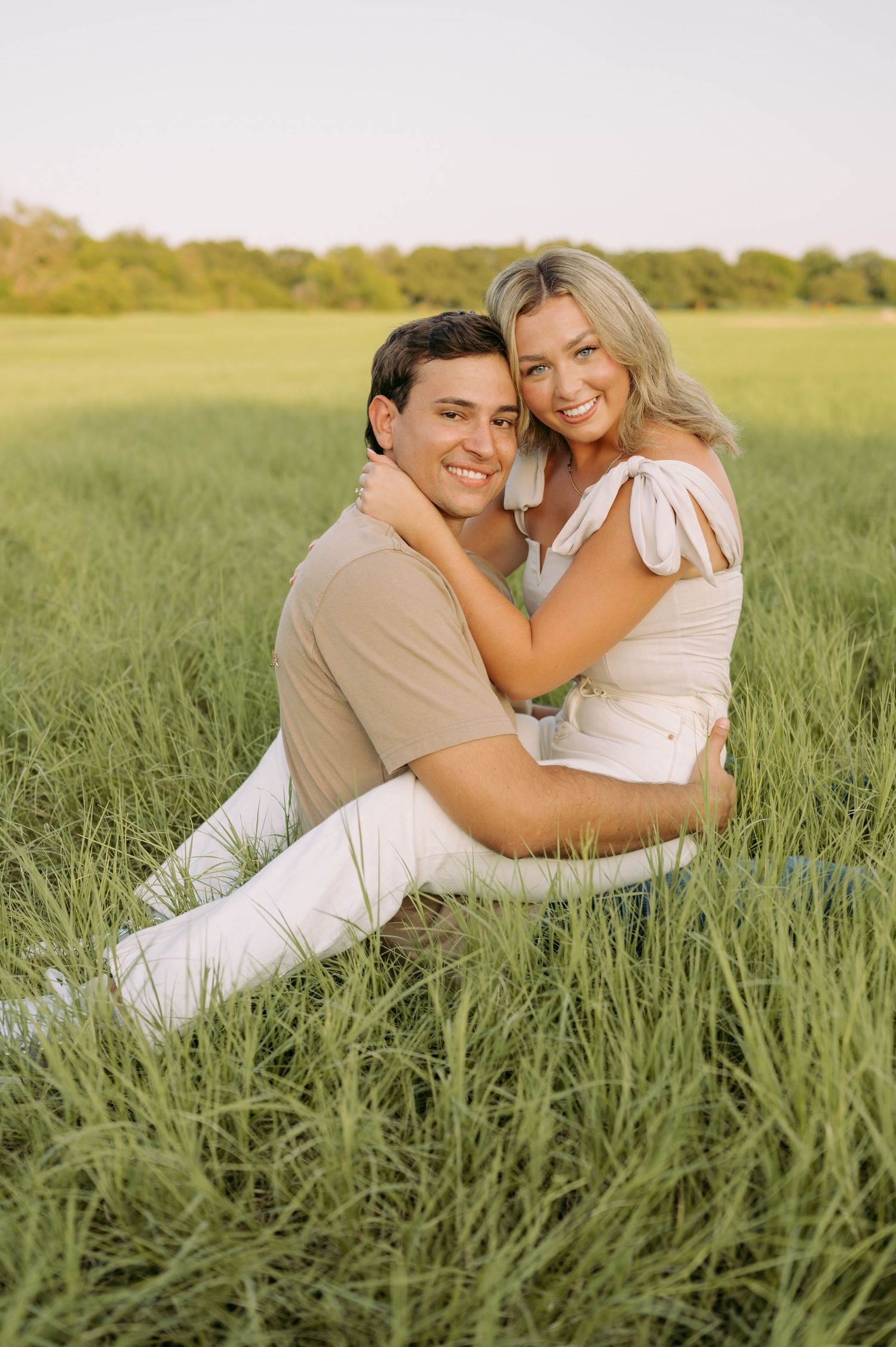 A couple sits together in a lush green field during golden hour, sharing an intimate embrace.