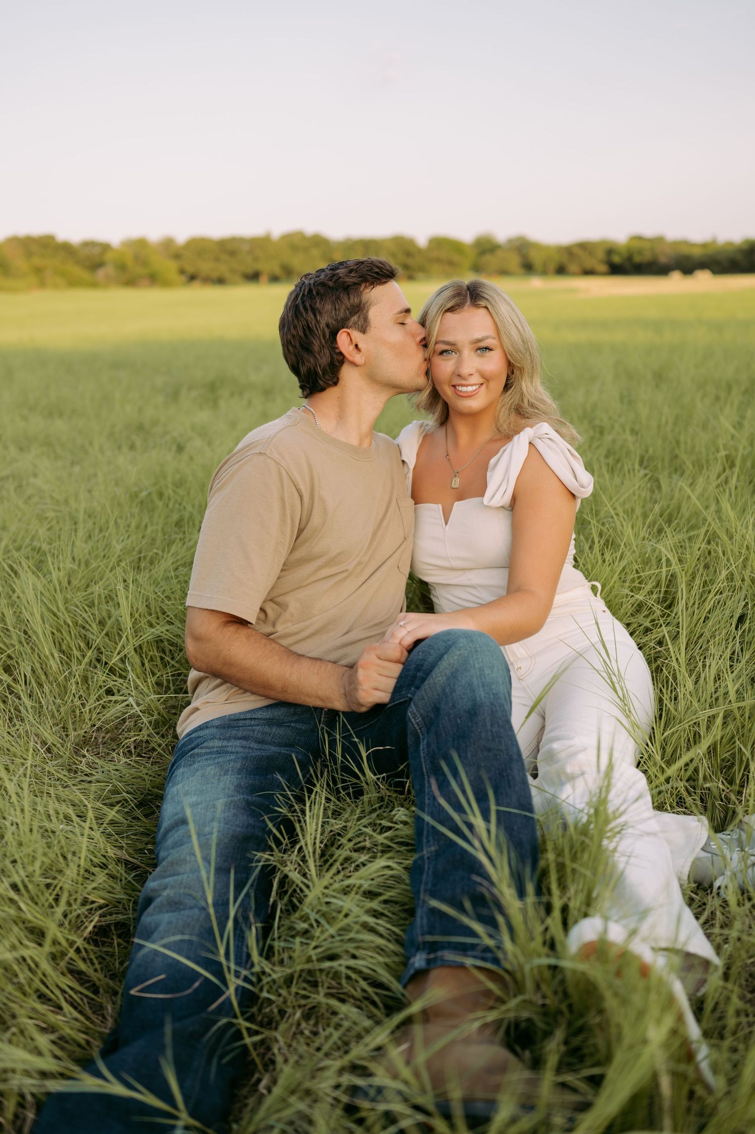 Two people share an intimate moment sitting together in a tall grass field at golden hour.