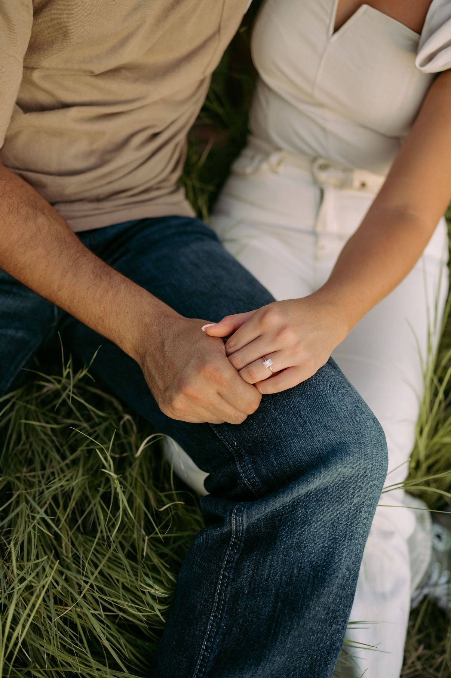 Two people sitting close together on grass, holding hands intimately, wearing casual jeans and light colored clothing.