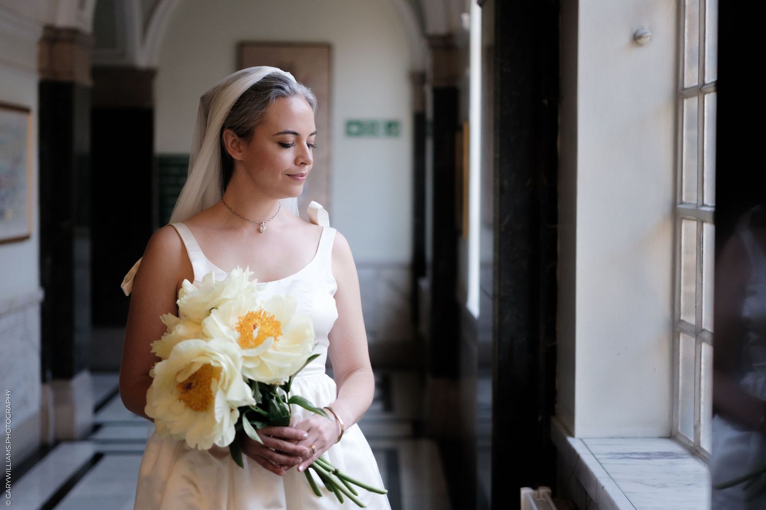 A person in a white dress stands in a hallway holding a bouquet of yellow daffodils by a window.
