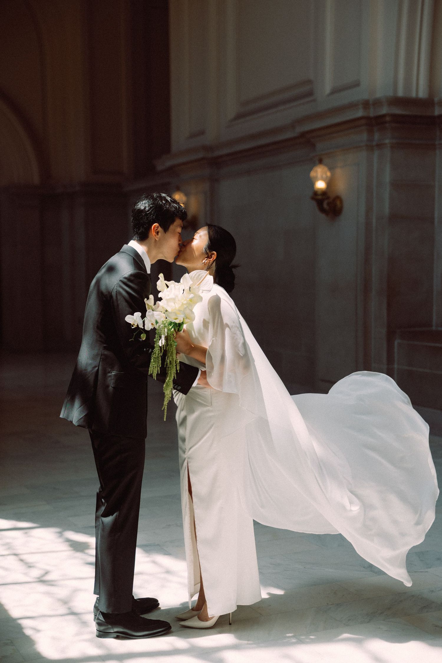 Wedding couple poses by ornate window in elegant venue with dramatic lighting.