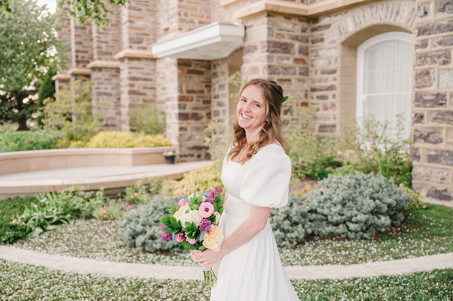 A person in a white wedding dress holds a colorful spring bouquet while standing outside a stone church building.