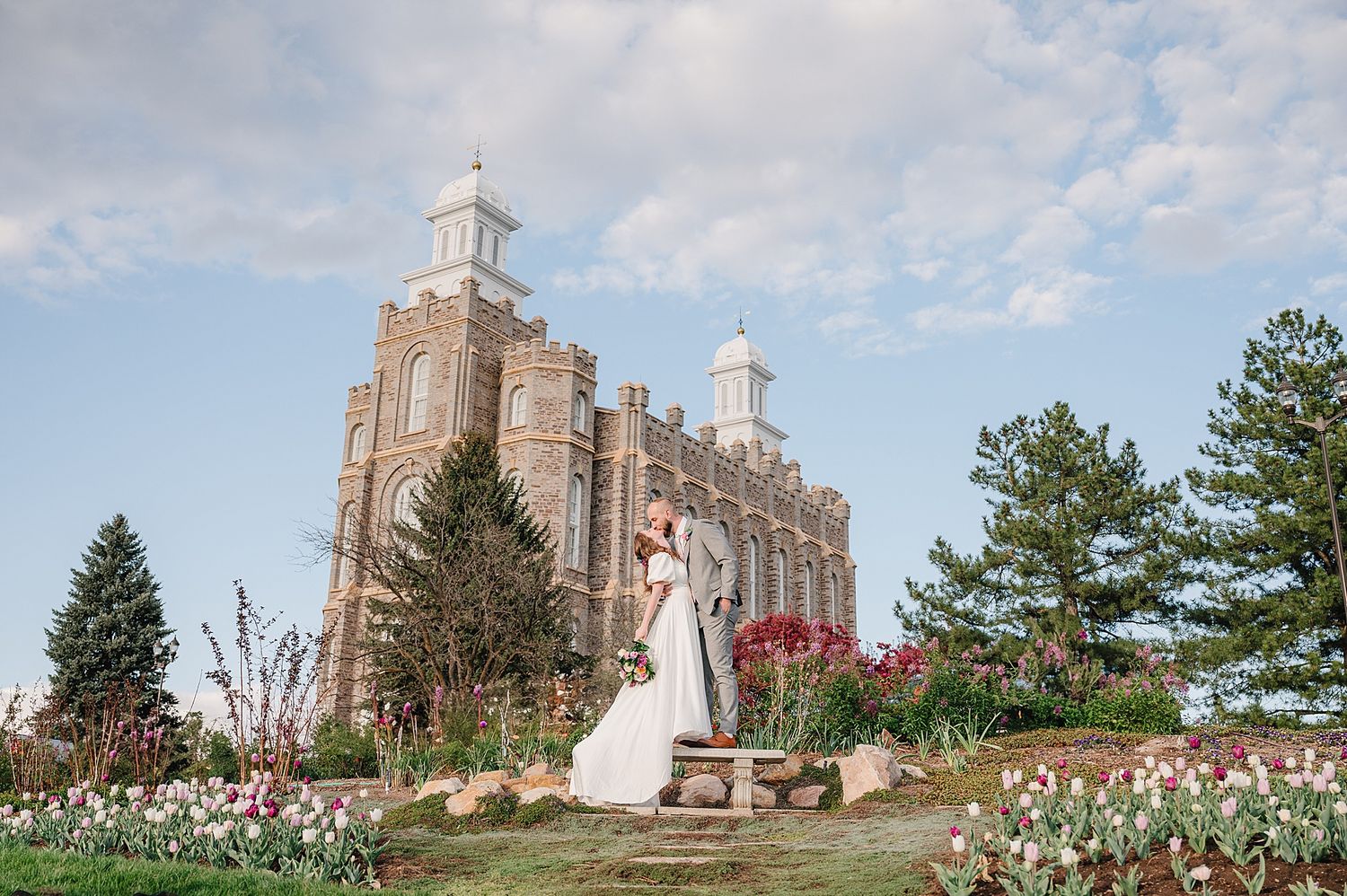 A couple stands in front of a majestic brick church with two spires, surrounded by blooming spring flowers in the garden.