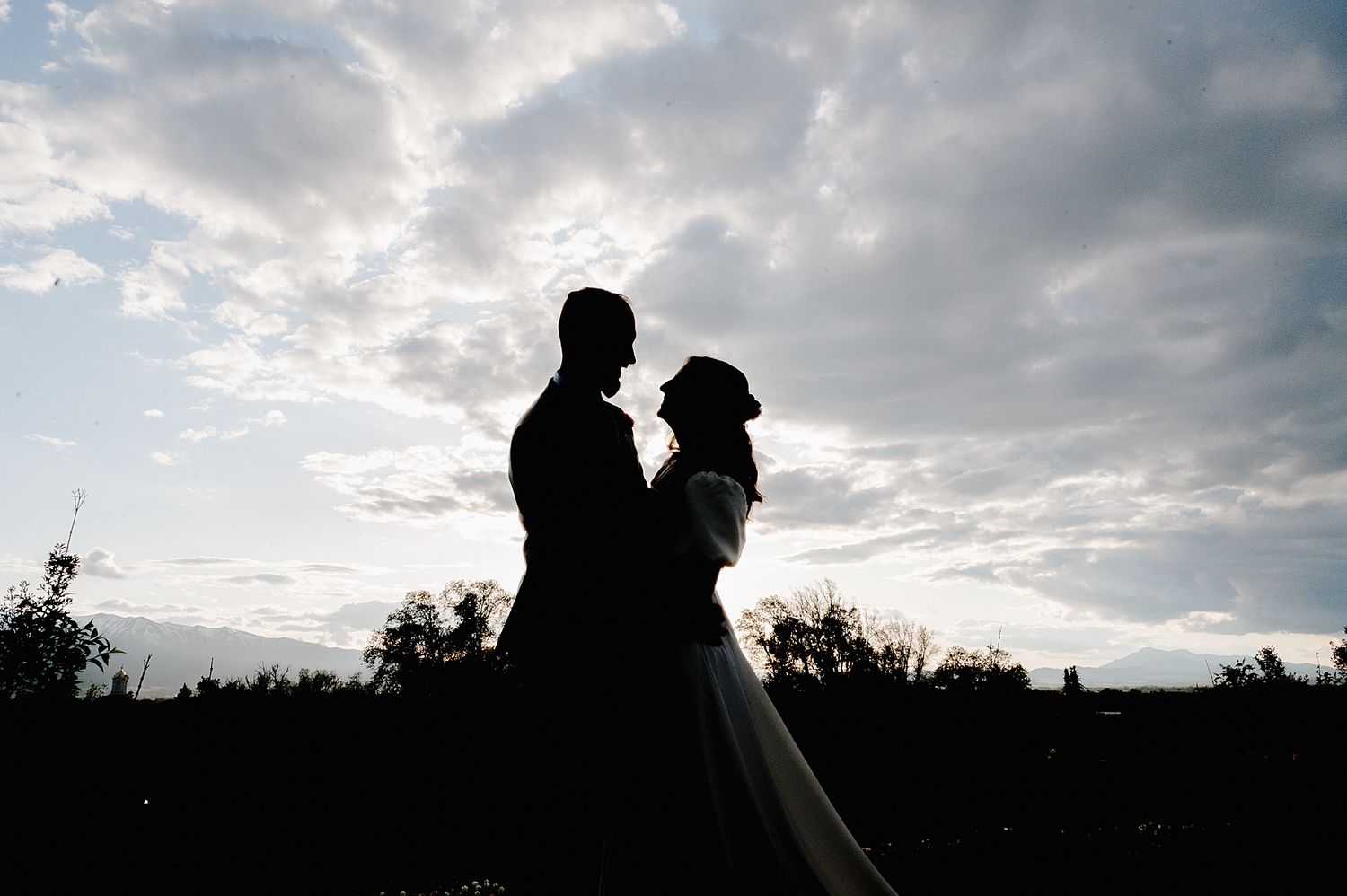 Silhouetted couple embrace against dramatic cloudy sky during sunset wedding photoshoot in nature.