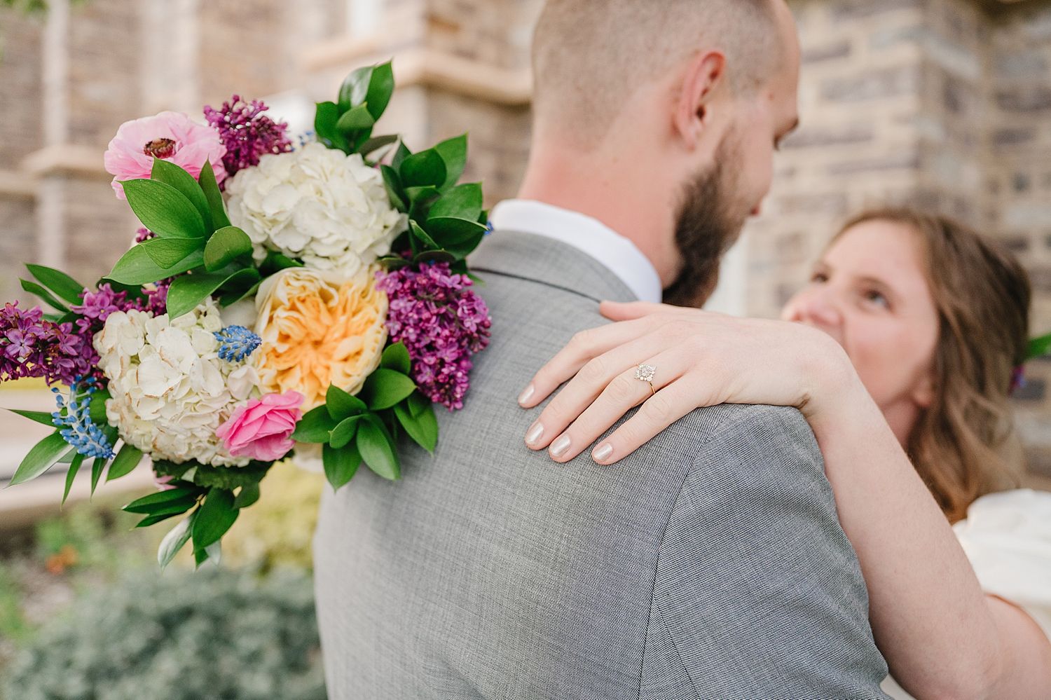 A beautiful wedding bouquet with white hydrangeas, pink roses and purple lilacs is held against a grey suit.
