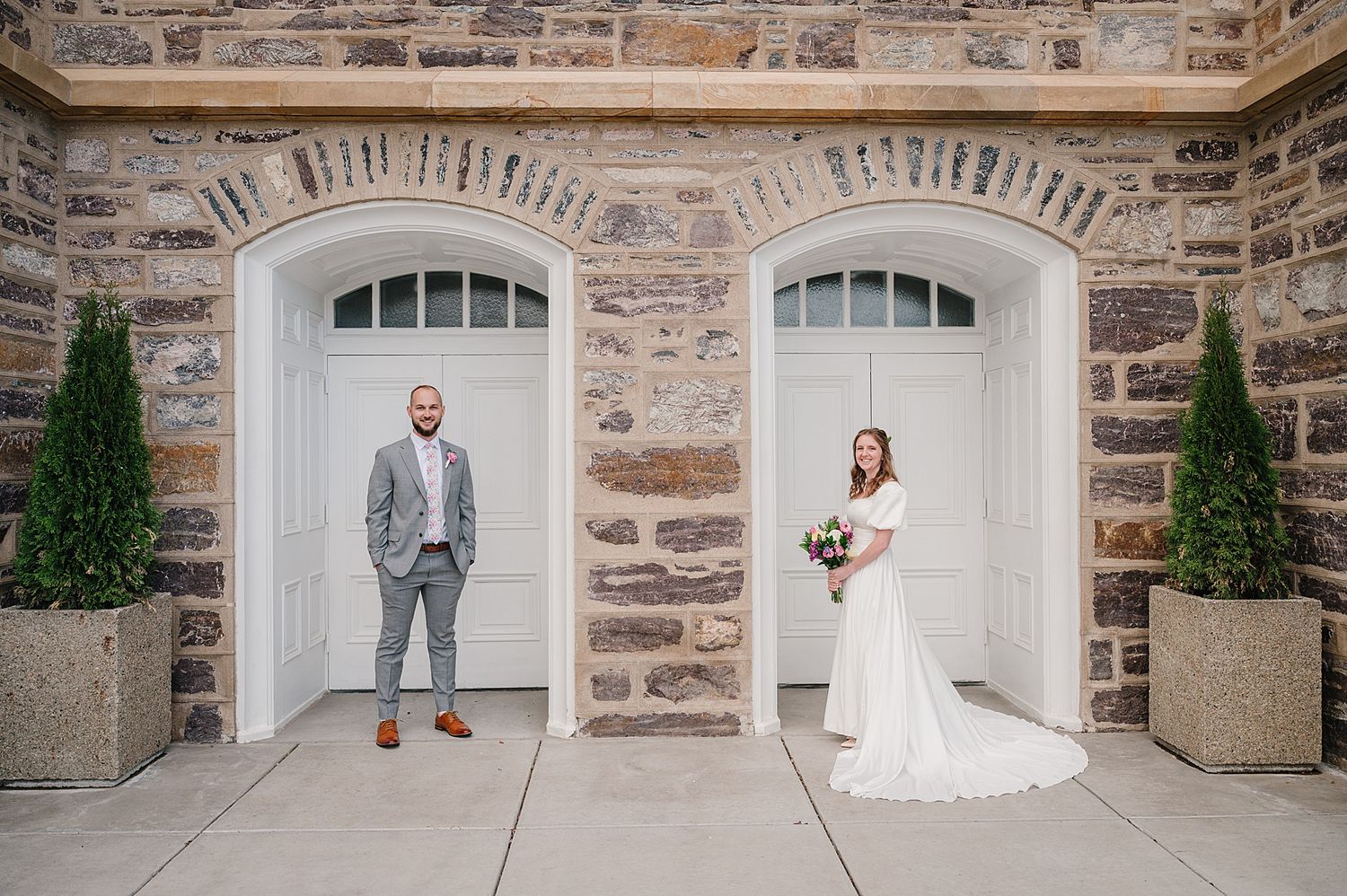 Two figures stand elegantly in front of grand white doorways with stone walls and potted topiary plants on either side.