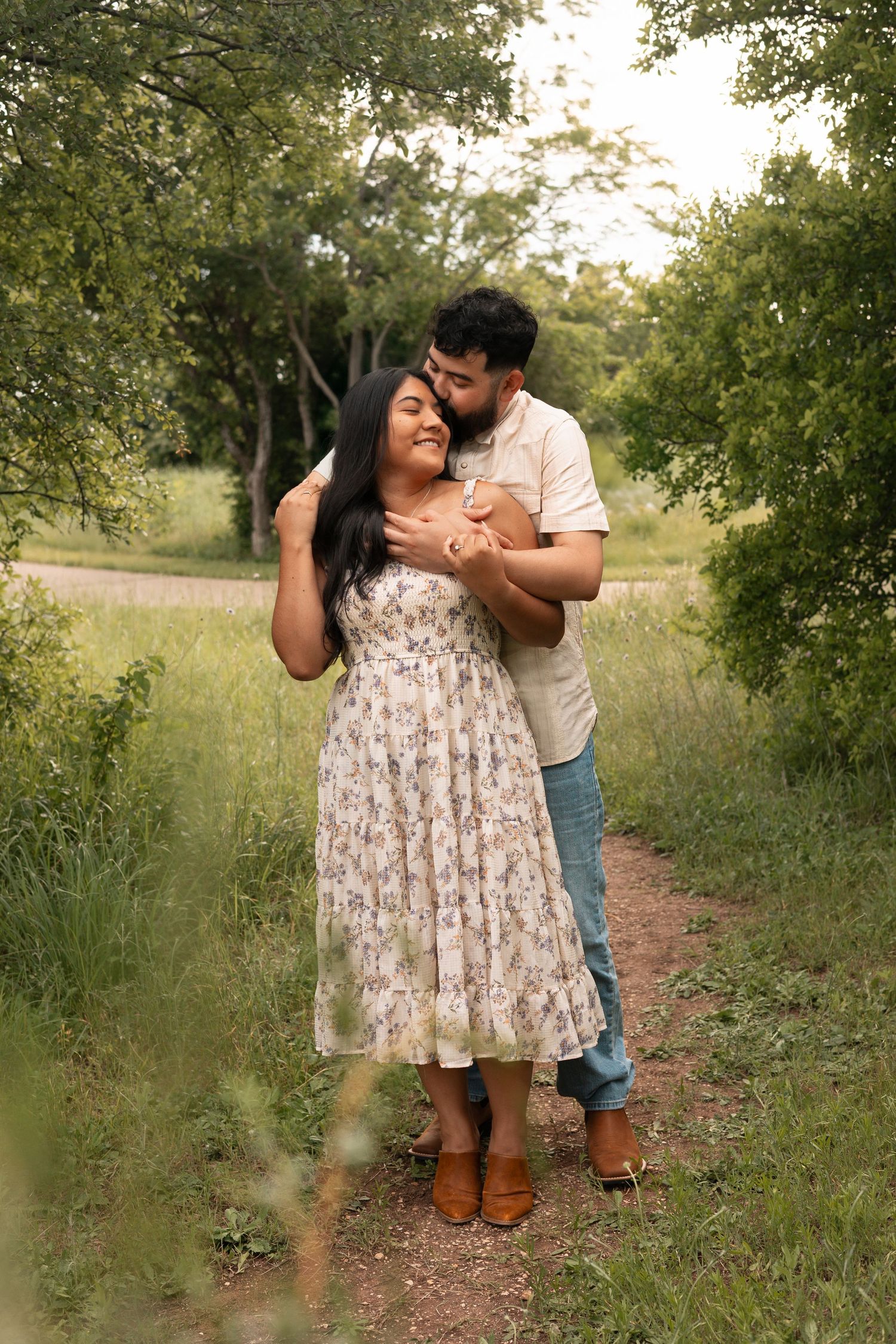 A romantic couple embraces on a nature trail surrounded by lush greenery and trees at golden hour.