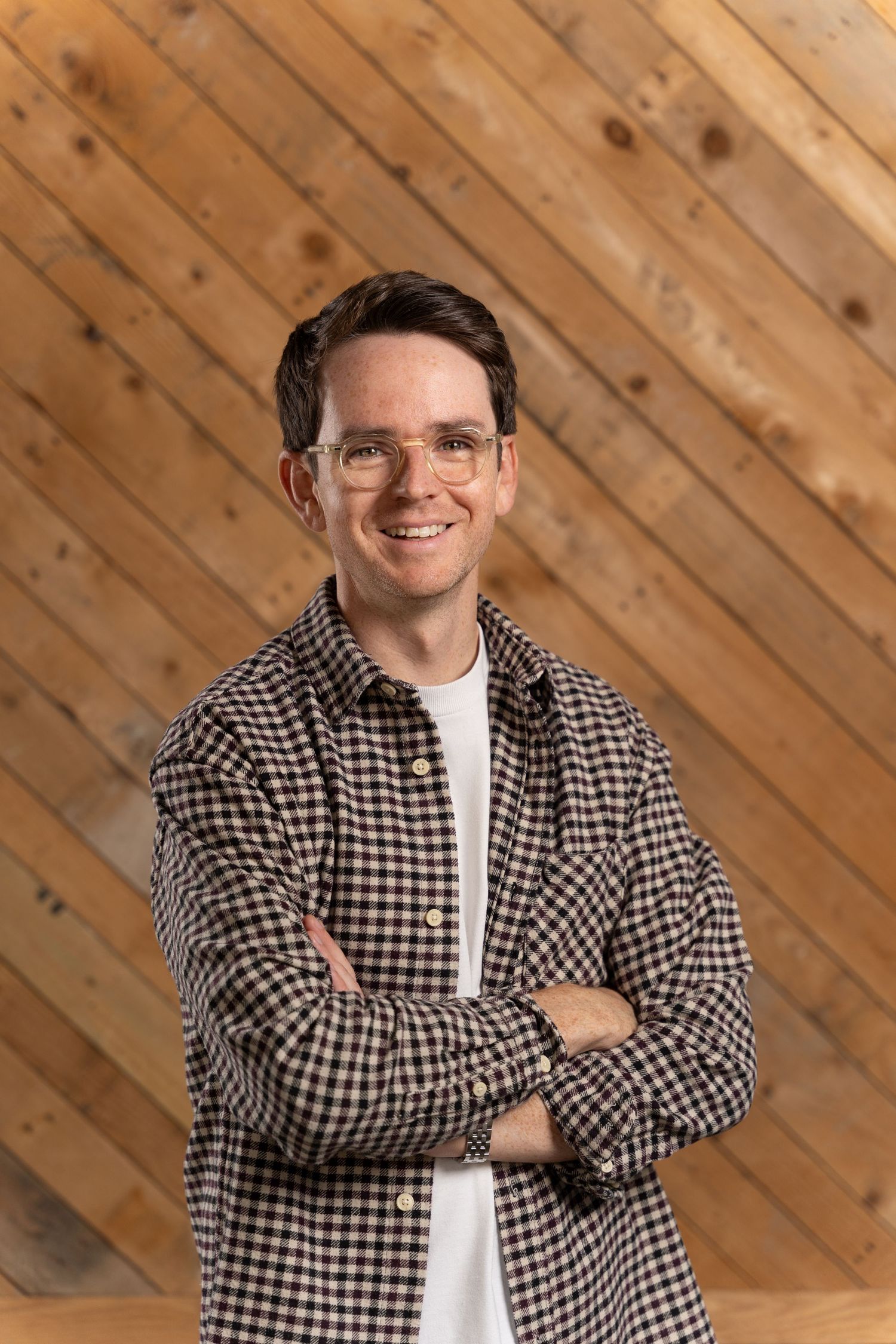 A person wearing glasses and a checkered shirt poses with crossed arms against a wooden paneled wall.