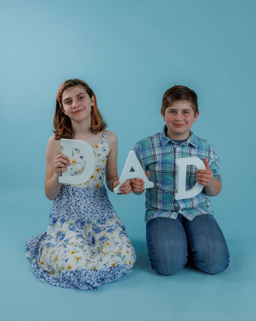 Two people kneel on blue backdrop holding decorative 'DAD' letters while wearing a floral dress and plaid shirt.