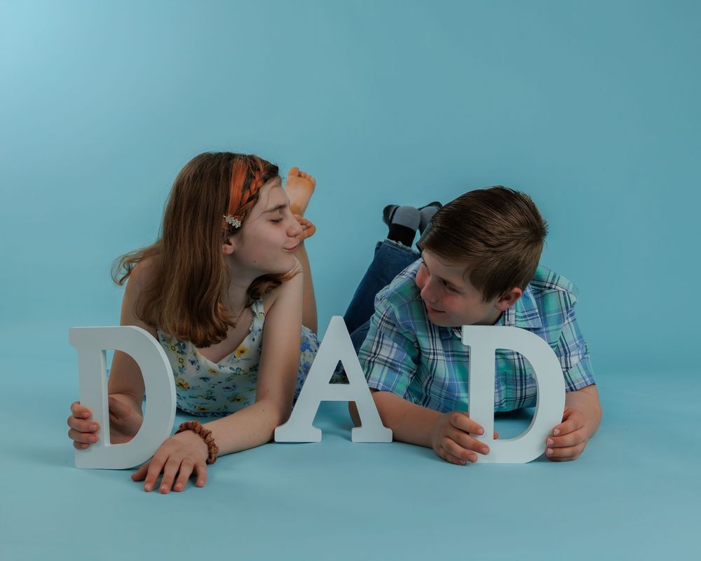 Two children lie on the floor holding large white letters that spell DAD against a turquoise background.