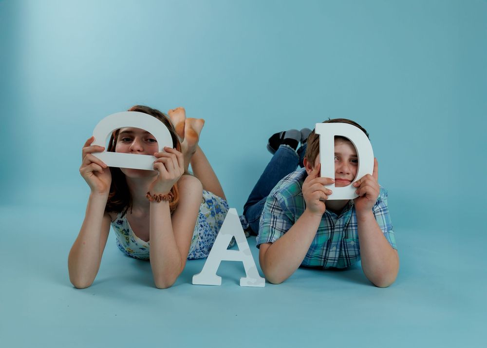 Two people lying on blue background holding up white letters that spell DAD in a playful pose.
