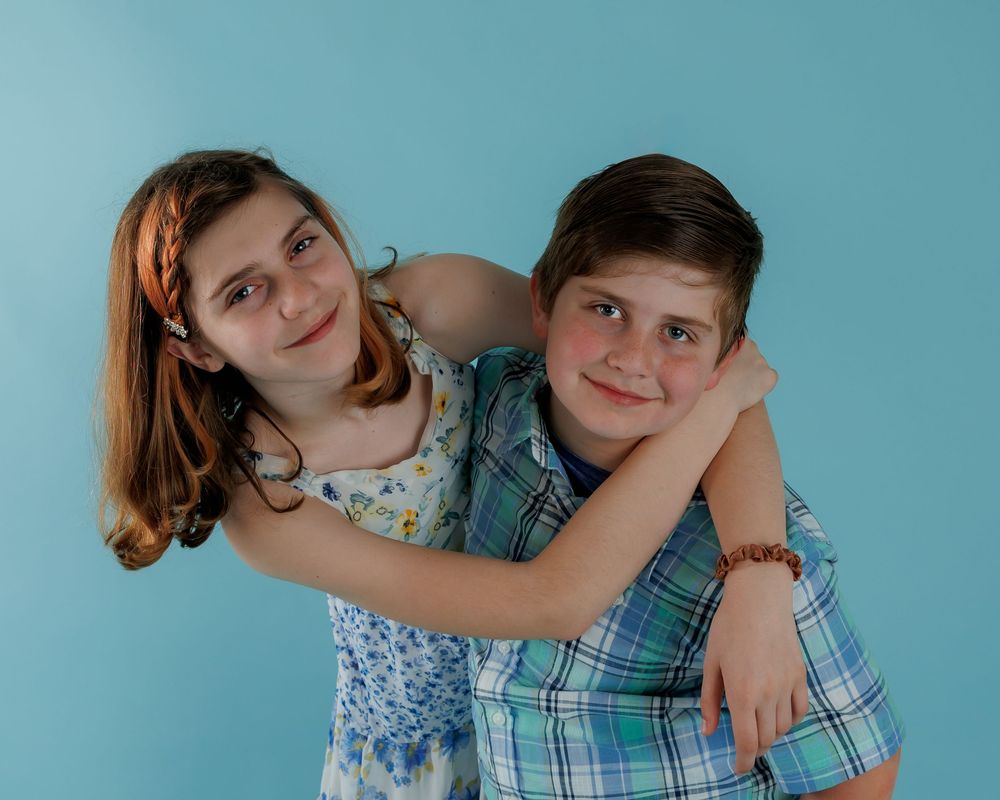 Two young siblings hugging and smiling against a blue background.