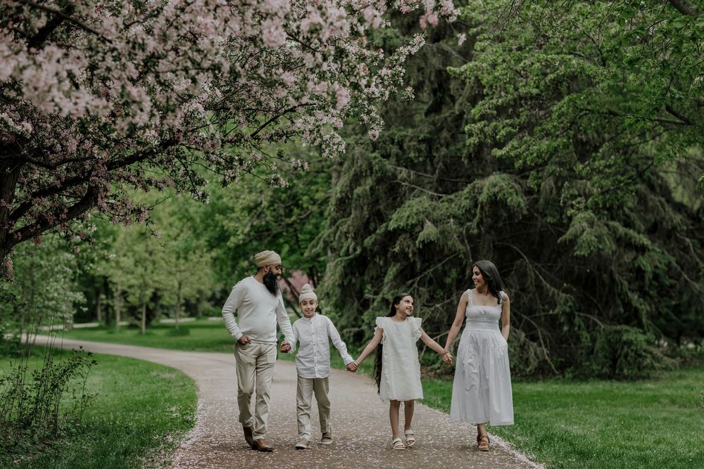 Family of four in white outfits holding hands on a garden path surrounded by blooming spring trees.