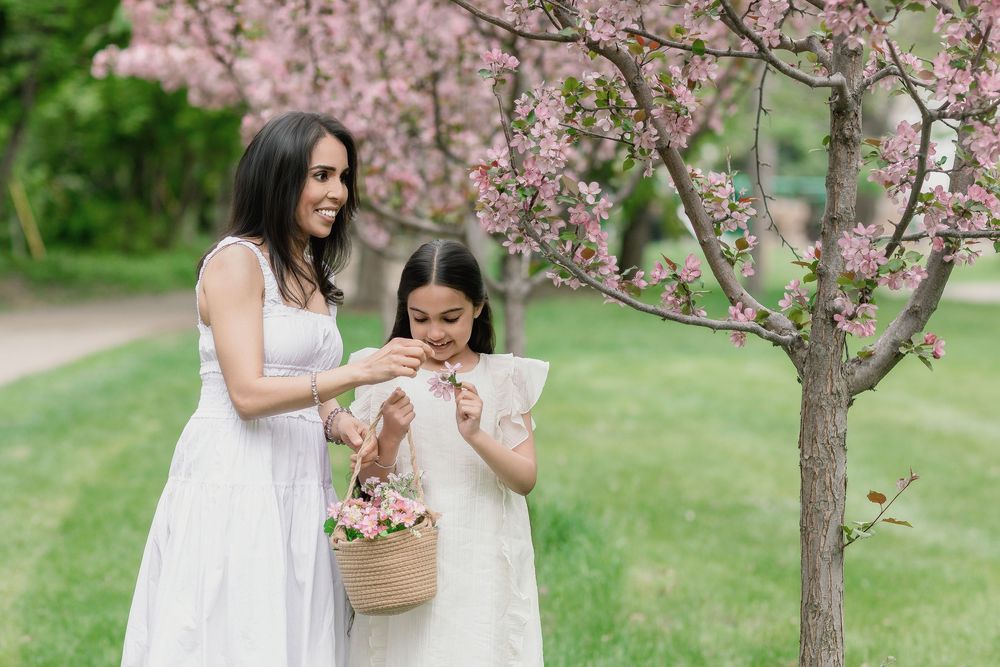 Mother and daughter in white dresses admiring pink flowers in a basket near a blooming cherry tree outdoors.