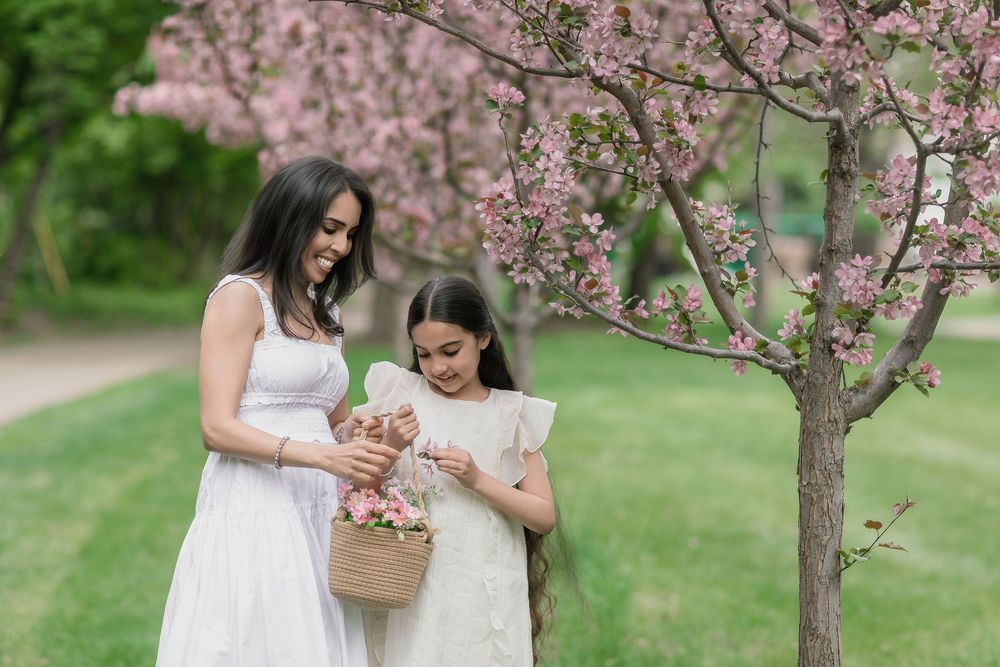 Mother and daughter in white dresses admiring pink flowers in a basket near a blossoming cherry tree in a park.