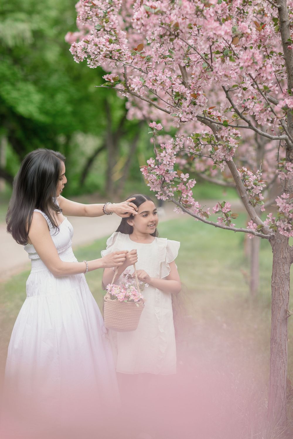 Mother in white dress fixes daughter's hair near pink cherry blossom tree, both wearing white dresses, holding flower basket.