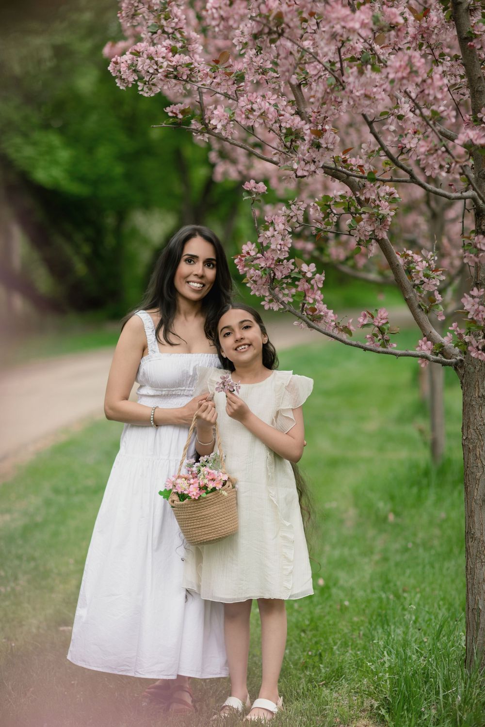 A mother and daughter in white dresses pose by a pink flowering tree holding a basket of flowers.